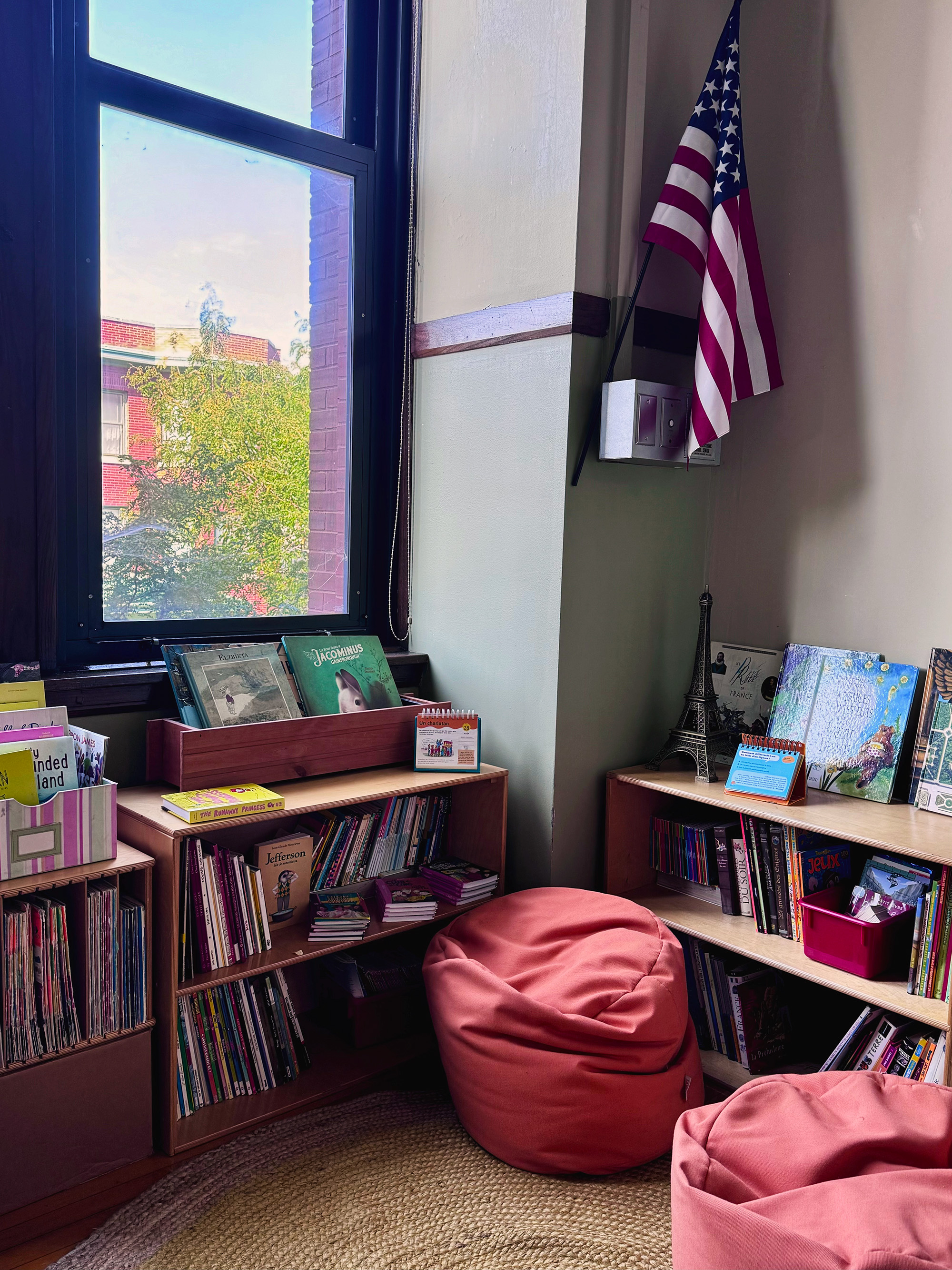 A view of the corner of a classroom shows an American flag, two pink beanbag chairs, and a set of bookshelves containing multi-lingual books