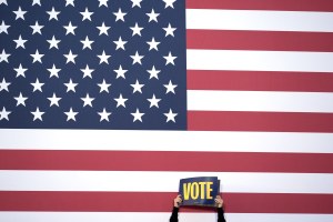 a person holds a sign that says vote in front of an American flag