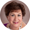 Ellen Shanley is pictured from the shoulders up, smiling into the camera wearing a purple shirt and white and gold necklace and matching earrings.