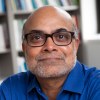 Prabhu Pingali is pictured from the shoulders up wearing a blue button-down shirt and smiling into the camera as he sits in a library.