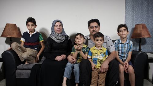 A family of 2 parents and 4 children of various ages sit on a couch