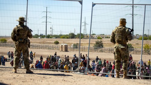 Migrants wait in line adjacent to the border fence under the watch of the Texas National Guard 