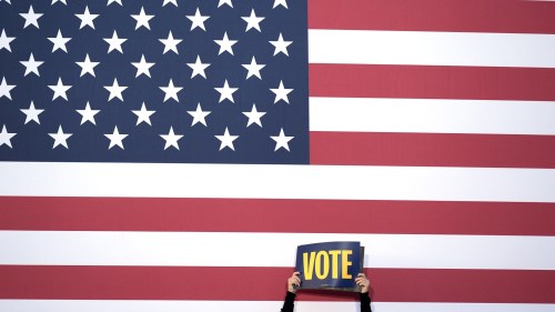 a person holds a sign that says vote in front of an American flag