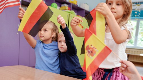 Small children at a classroom desk wave flags from the U.S., Germany, and Spain