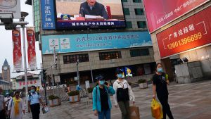 People hold shopping bags as they walk under a giant screen showing a news footage of Chinese President Xi Jinping, at a shopping area in Beijing