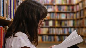 A young girl reads a book, with bookshelves of other books in the background