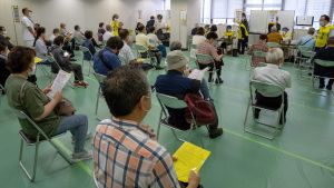 People sit in chairs in a large room, six feet apart