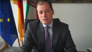 Screenshot of the virtual symposium, of a man at a desk in front of flags