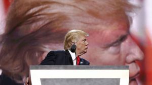 Donald Trump standing at a podium, with a TV screen behind him