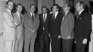 A black and white photo of seven men standing in a semicircle. 