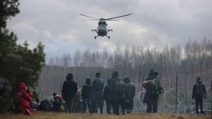 People stand in front of a barbed wire fence while a helicopter flies in the background