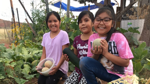 Three young girls kneel in a garden, holding vegetables and fruits as they smile into the camera.