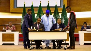 2 men sit at a desk while 3 others stand between them, with flags in the background