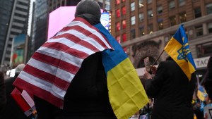 Person seen from behind with an American flag and Ukraine flag draped over their shoulders, grey beanie on their head.