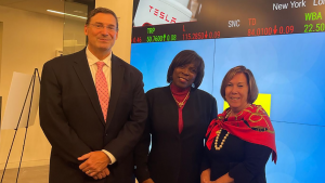 Ertharin Cousins stands in the middle of two people, all smiling into the camera. Behind them is a large screen displaying the news.