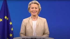 Ursula von der Leyen stands in front of blue screen with European Union flag to the left.