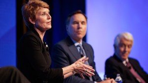 A person talks on stage during an event while two other people listen in the background