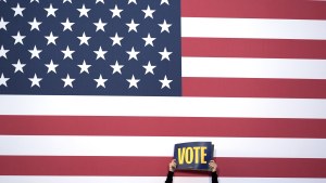 a person holds a sign that says vote in front of an American flag