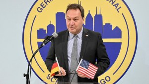 A man stands at a podium with German and American flags on it, speaking in front of a blue and yellow circular logo on a wall